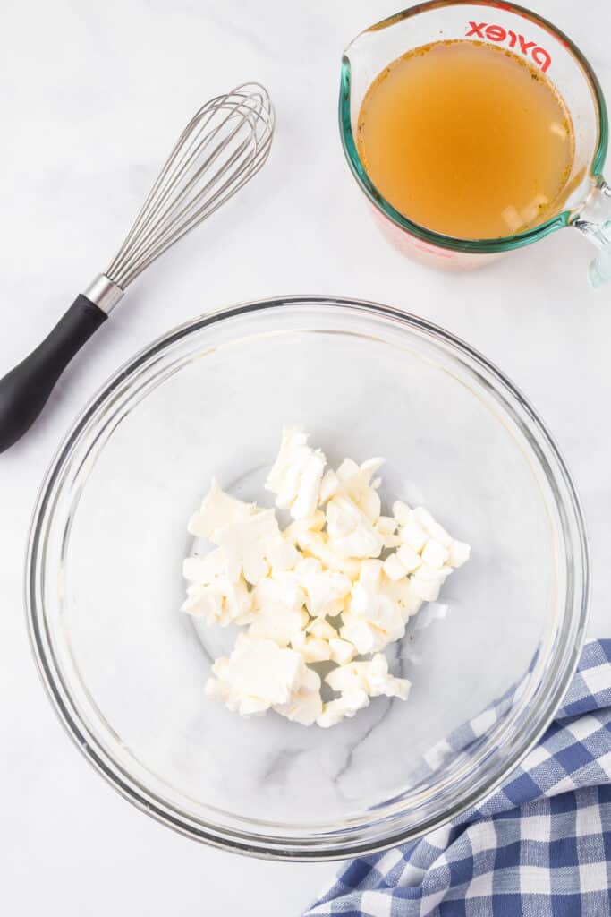 Pieces of cream cheese in a large mixing bowl with a whis and a measuring cup full of broth on the counter nearby from overhead.
