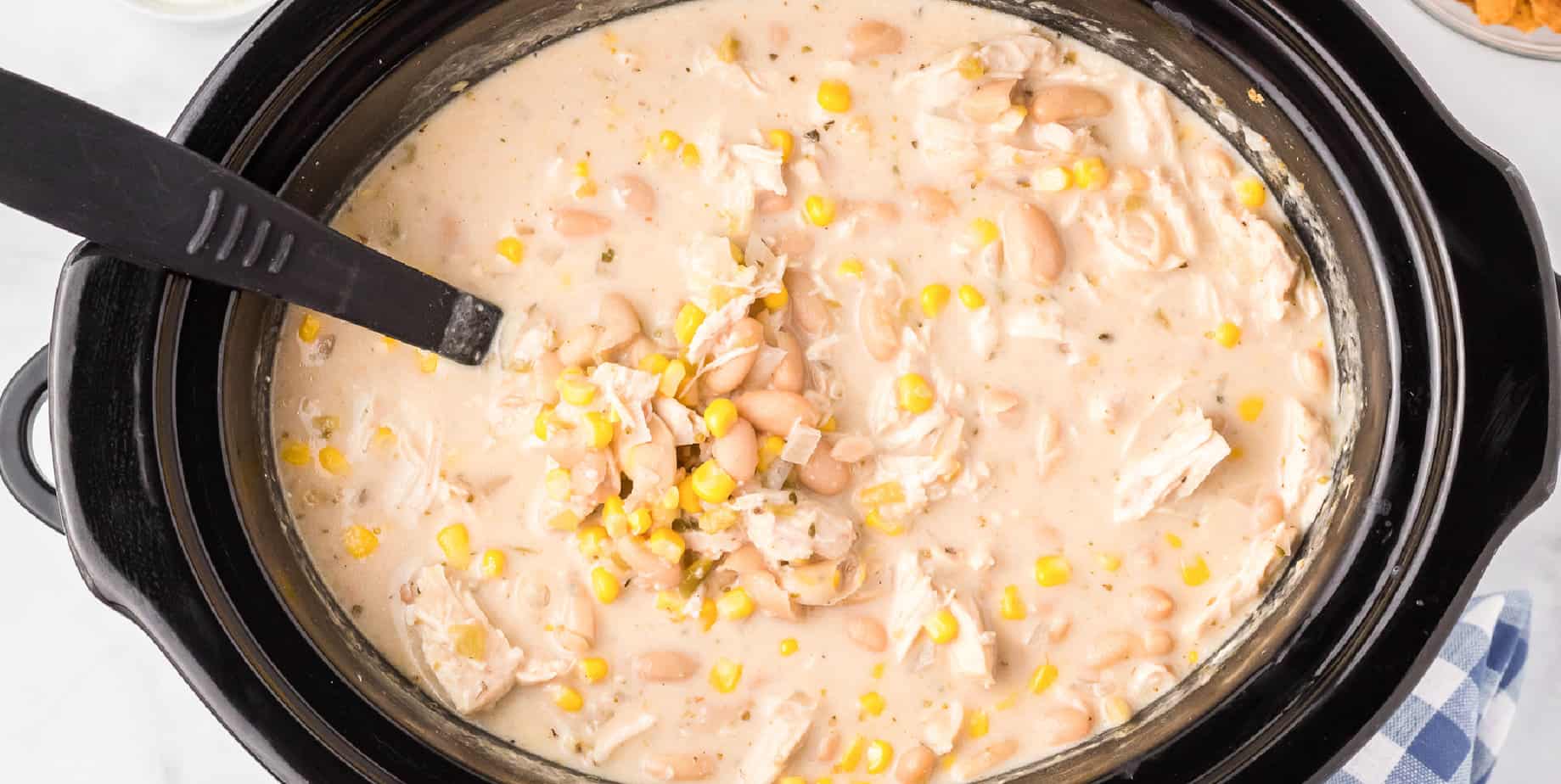 Overhead wide view of creamy chicken toritlla soup with corn in a slow cooker from above with a ladle close up.