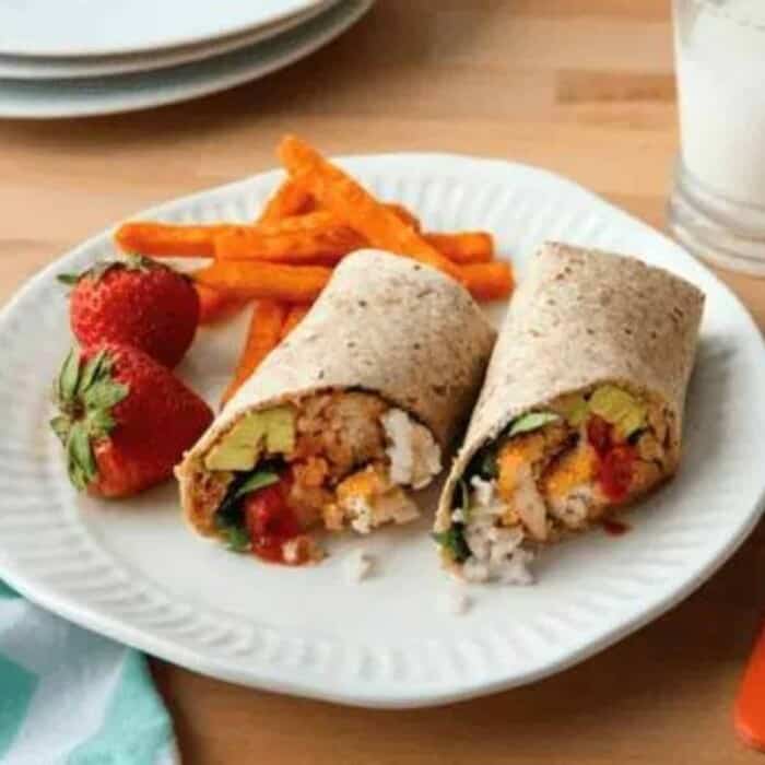 wrap with chicken nuggets, cooked rice, shredded cheese, and avocados, cut in half diagonally on a white paper plate with sweet potato fries and strawberries.