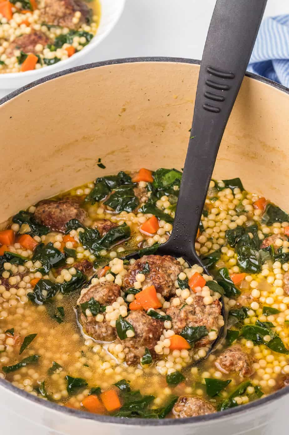 Close up of Italian wedding soup being scooped by a ladle from a large pot with a bowl full nearby on the counter.
