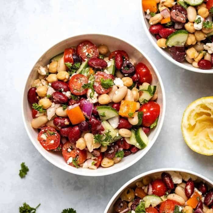 three white bowls with kidney beans, halved grape tomatoes, garbanzo beans, white beans, chopped red onion and zucchini.