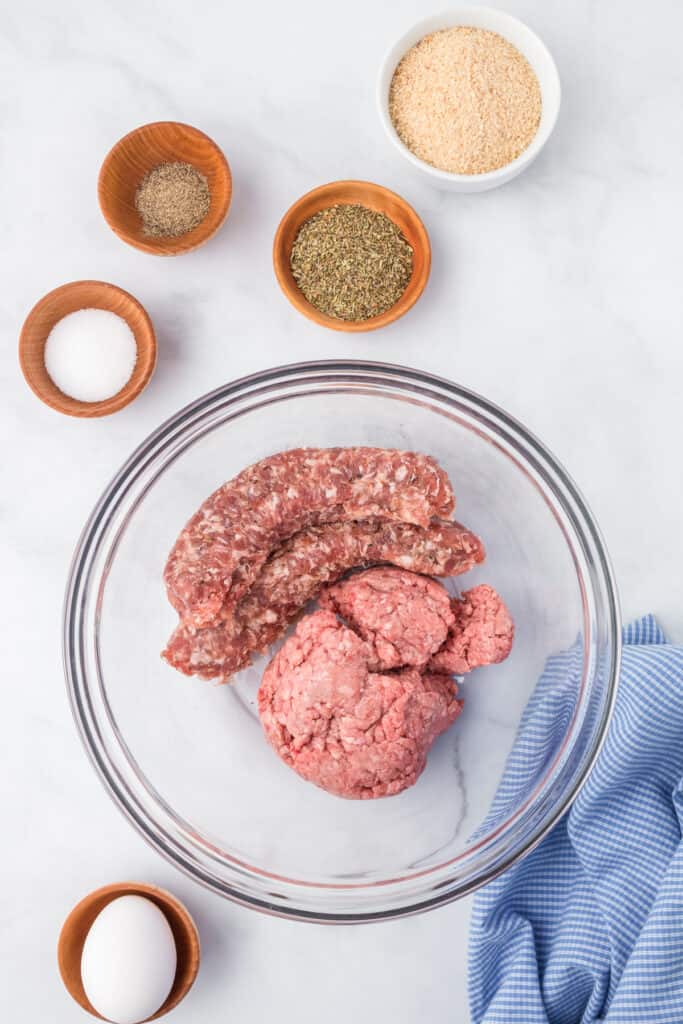 Mixing together ground beef and sausage links in a bowl on a counter from overhead with more spices on the counter nearby for italian wedding soup.