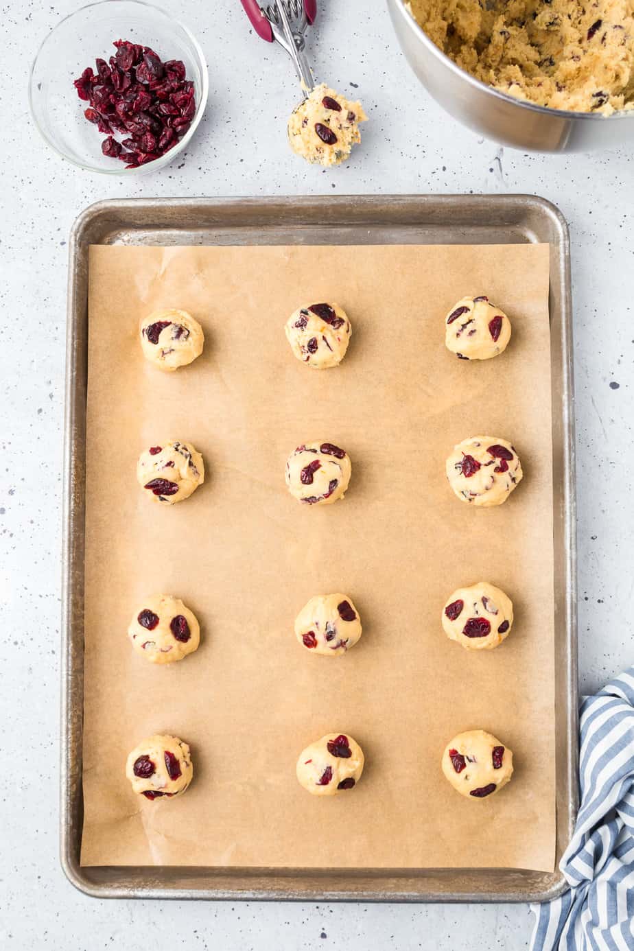Cooke dough balls space on a parchment lined baking pan with cranberries on top of each cookie dough ball on a counter form above.