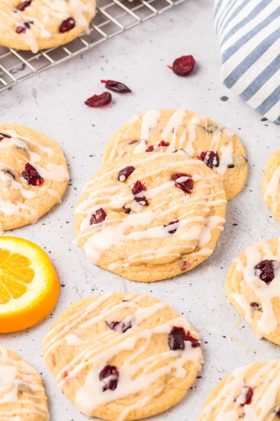 Glazed cranberry cookies piled on a counter with more cranberries and slices of orange nearby from the side.
