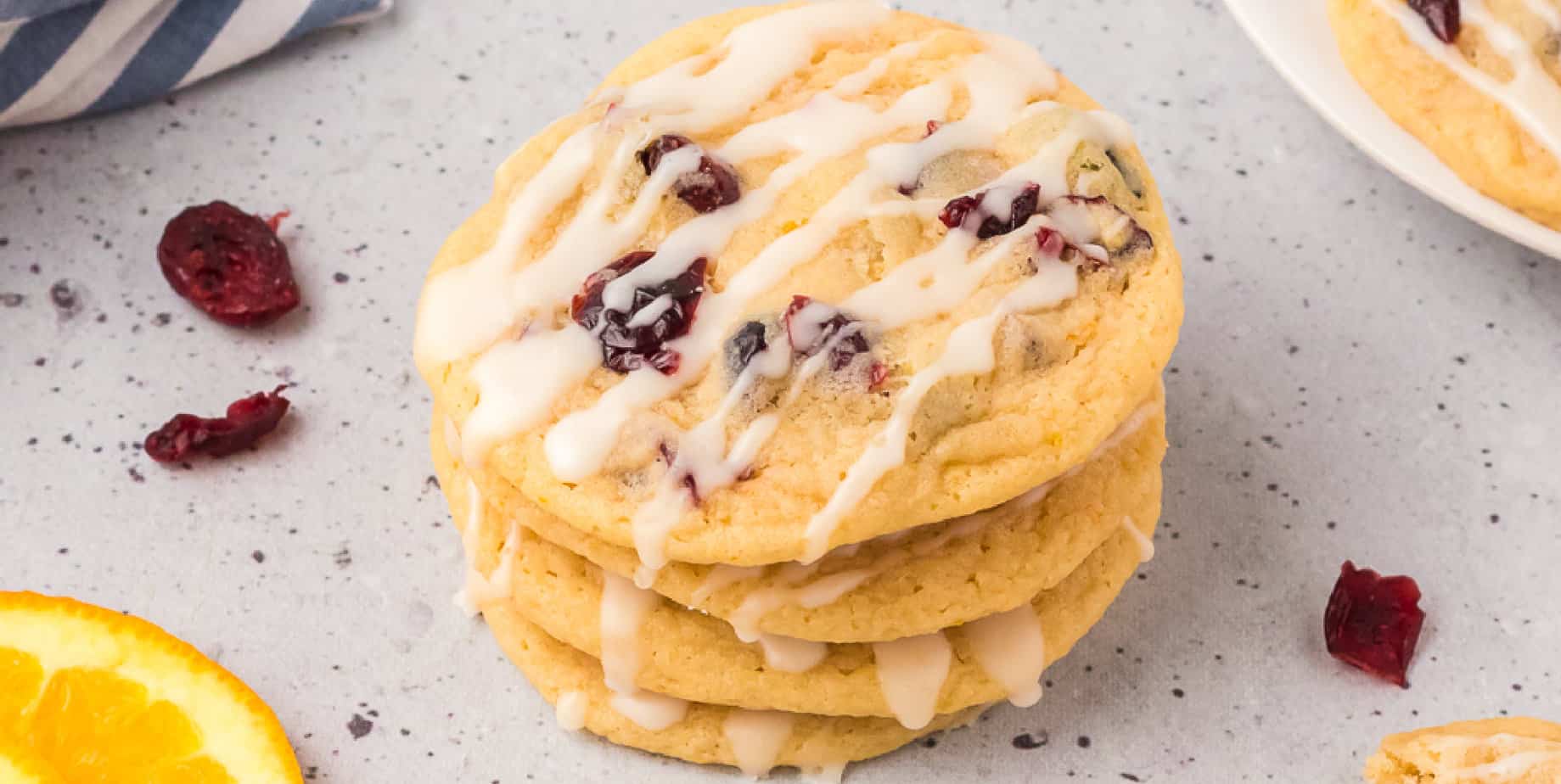 A stack of cookies on a counter with cranberries and glaze on top, with more cranberries and a slice of orange nearby on the counter.