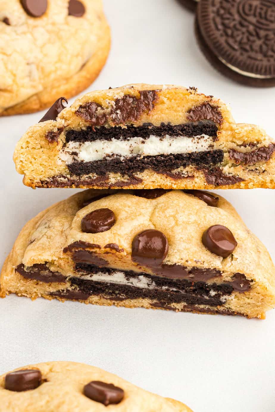 Close up of a chocolate chip cookie stuffed with an Oreo sliced in half up close on a counter with more chocolate chip and Oreo cookies nearby.