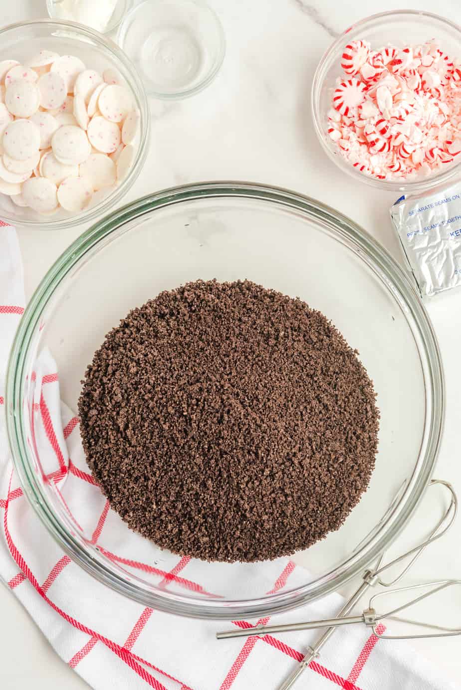 Chocolate cookie crumbs in a large bowl on a counter with peppermint extract, peppermints, and white chocolate peppermint wafers in bowls nearby.