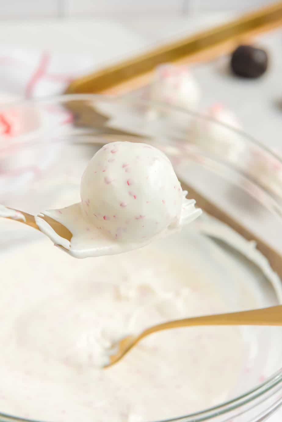 A white chocolate Oreo ball being lifted from a bowl of white chocolate by a fork with excess chocolate dripping back into the bowl from the side on a counter.