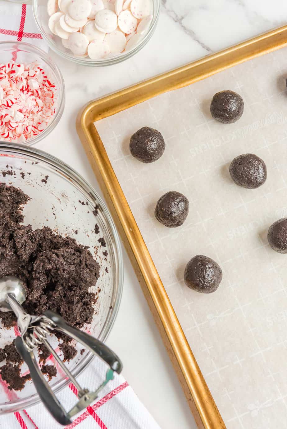 Chocolate dough being scooped with a cookie scoop into balls and place onto a parchment paper lined cookie sheet from overhead on a counter.