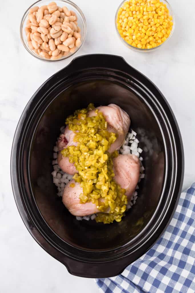 Raw chicken breast covered in green chilis with diced onions in the bottom of a slow cooker on a counter from overhead.