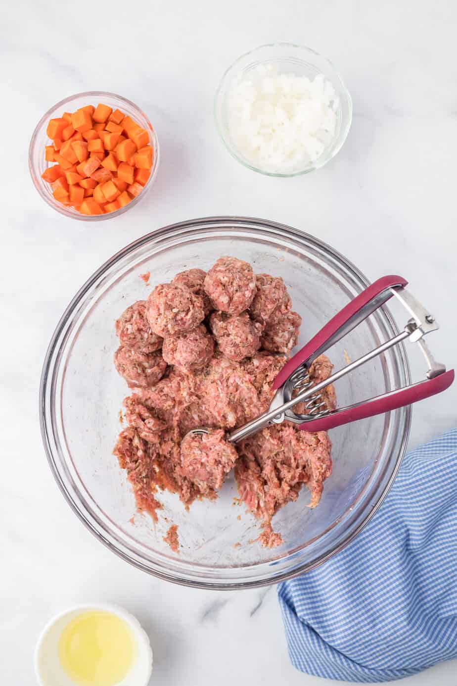 Scooping meatballs from a large glass bowl of mix on a counter from overhead with more ingredients nearby.