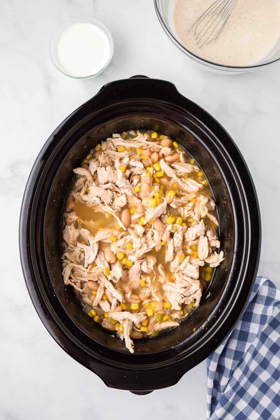 Shredded chicken in a slow cooker from overhead mixed into the soup from overhead with cream and a cream cheese mixture nearby on the counter.