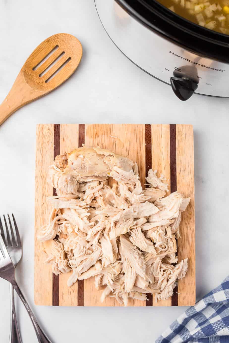 Chicken being shredded on a cutting board on a counter from overhead with a slow cooker, two forks and a wooden spoon nearby.