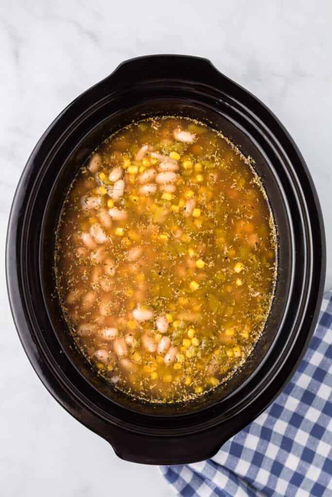 A slow cooker on the counter full of brown broth, white means, corn and chicken from overhead.
