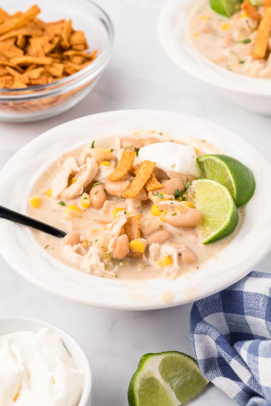 Creamy chicken chili in a bowl from the side on a counter with lime, tortilla shreds,, sour cream and a spoon in the bowl, and more toppings in bowls nearby.