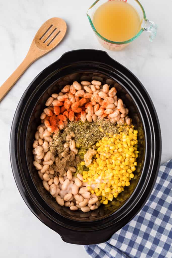 Slow cooker from overhead on a counter full of white means, chicken, green chilis, corn and spices with a measuring cup of broth and a wooden spoon nearby from overhead.