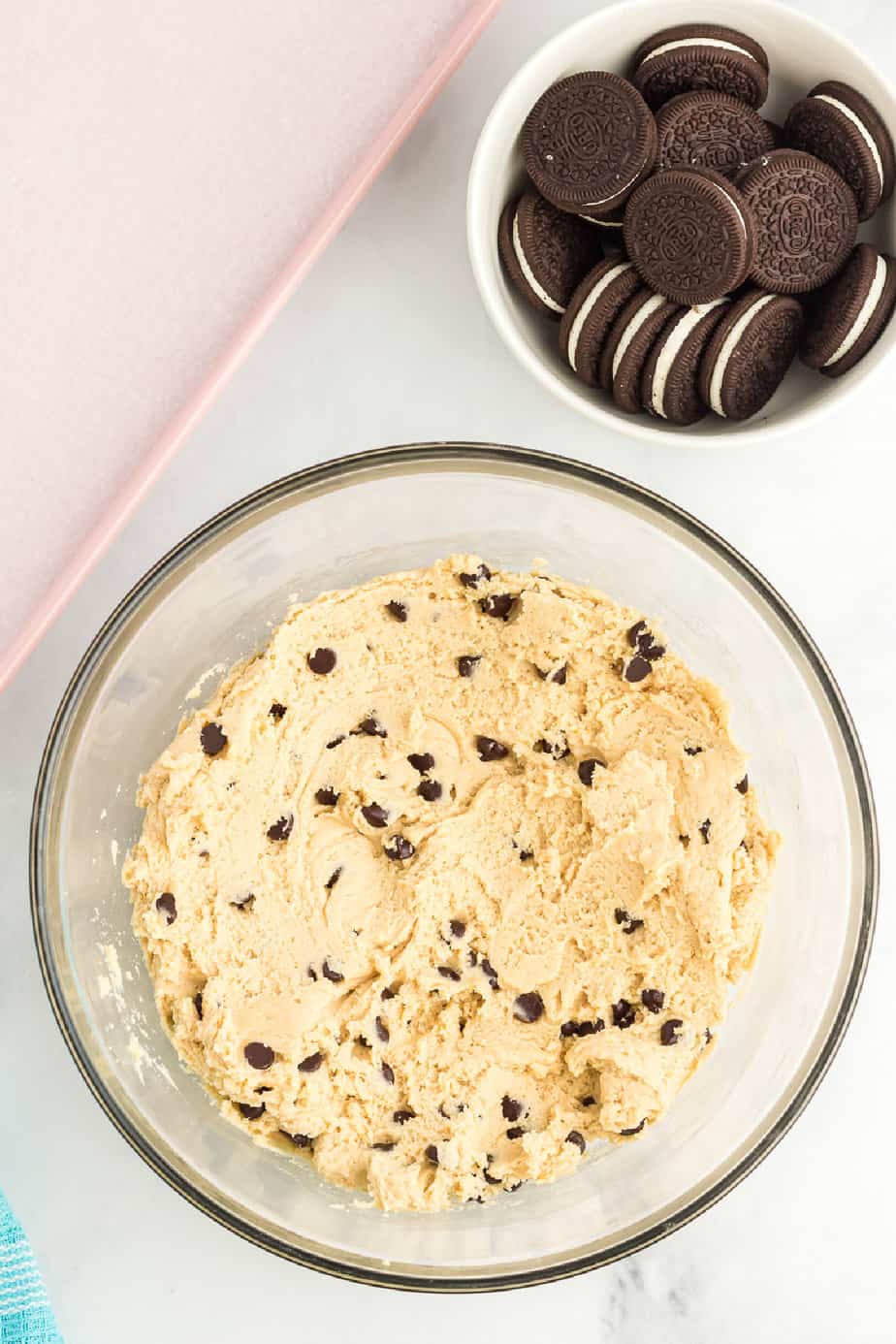 Cookie dough mixed with chocolate chips in a large bowl on a counter from overhead with a bowl of oreos and a pink baking sheet on the counter nearby.
