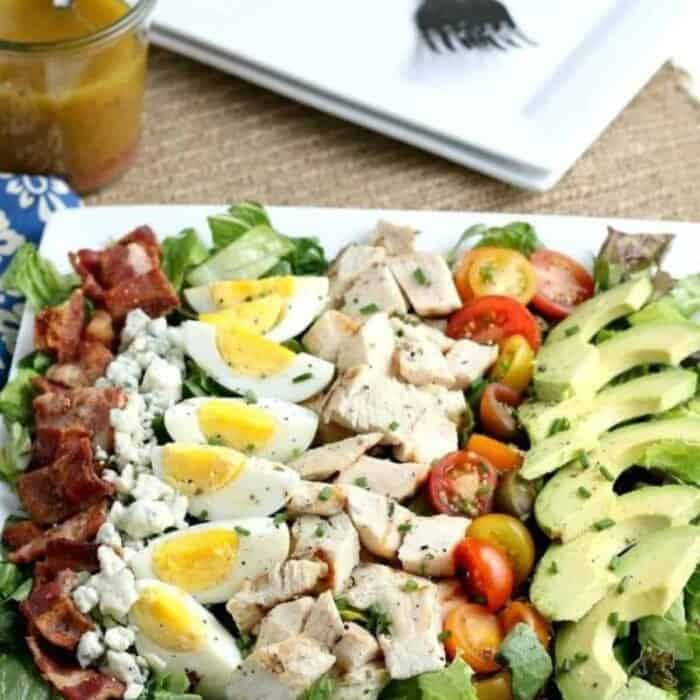 chopped cooked bacon, feta cheese crumbles, quartered hard boiled eggs, chunks of cooked chicken, halved grape tomatoes, and slices of avocado over a bed of romaine lettuce on a square white plate.