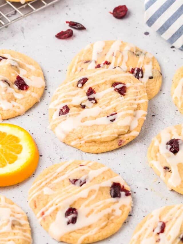 Glazed cranberry cookies piled on a counter with more cranberries and slices of orange nearby from the side.