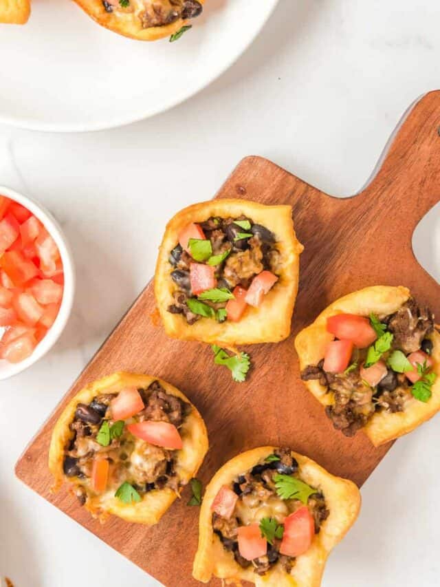 Four taco appetizers on a cutting board to serve with more on a plate nearby from overhead on a counter.