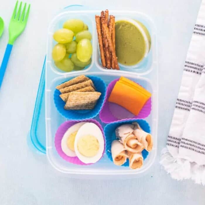 green grapes, small pretzel sticks next to a single serving container of guacamole, shredded wheat crackers in a blue silicone muffin liner, 2 square slices of cheddar cheese in a pink silicone muffin liner, a boiled egg cut in half lengthwise in a pink silicone muffin liner, rolled pieces of deli meat in a blue muffin liner, all in a plastic container with two small and one large compartment.