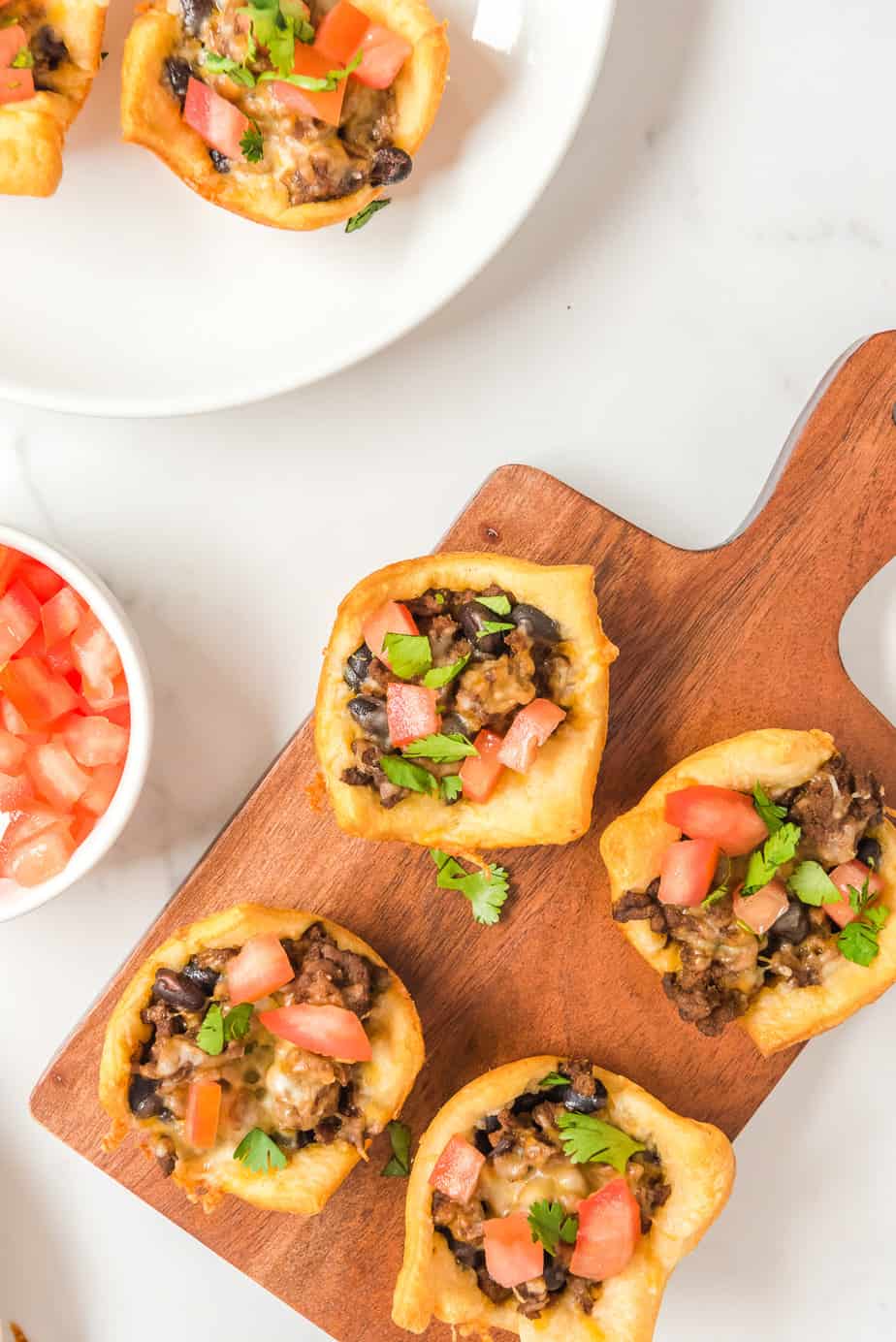 Four taco appetizers on a cutting board to serve with more on a plate nearby from overhead on a counter.
