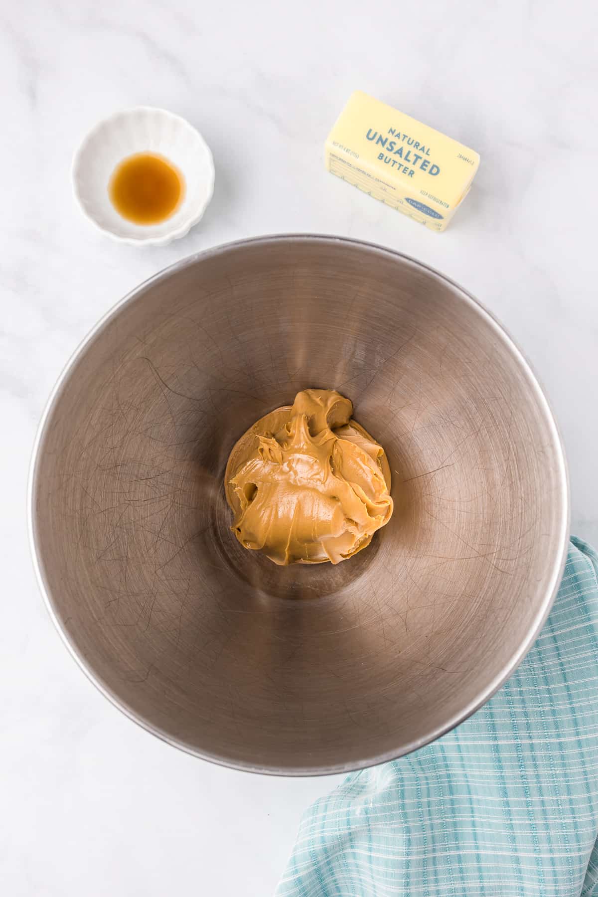 Peanut butter in a large mixing bowl on a counter from overhead with butter and vanilla nearby on the counter.