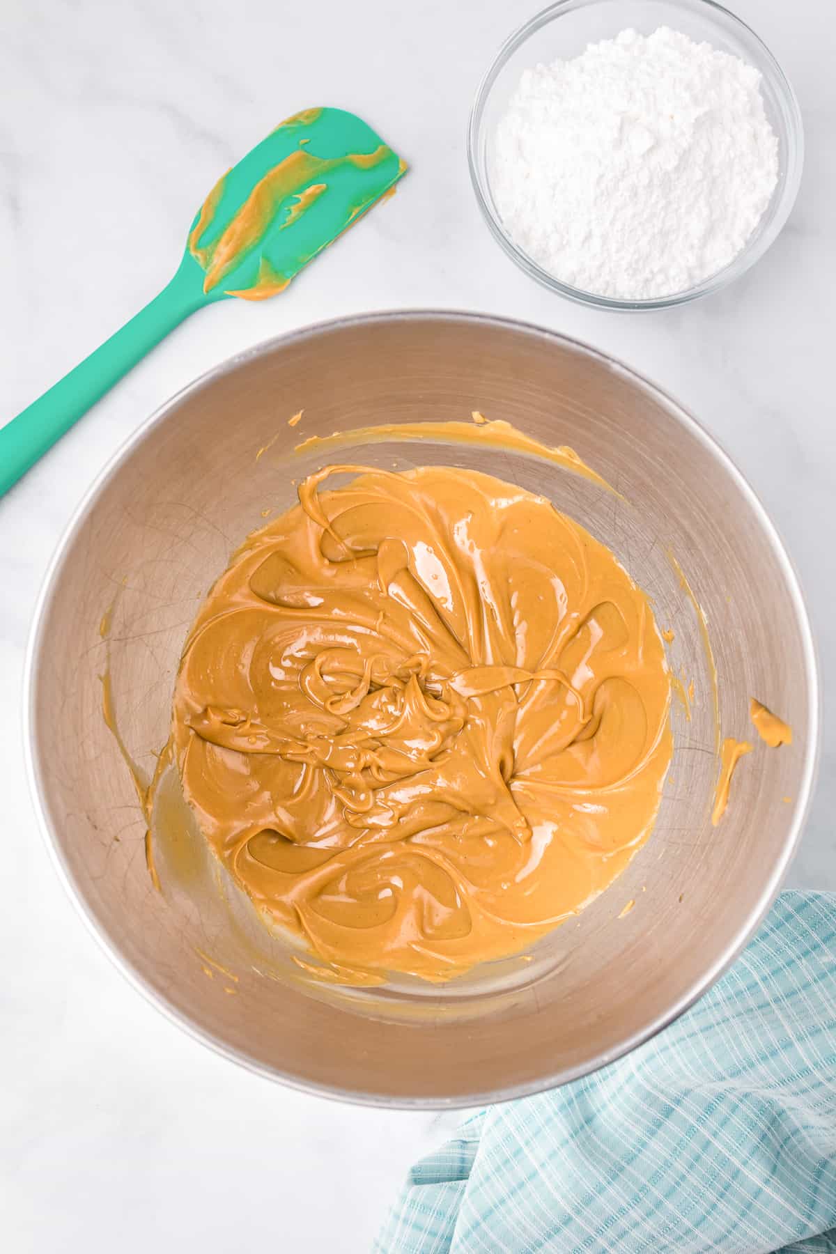 A peanut butter mixture in a large mixing bowl from above on a counter with a peanut butter doughcovered spatula and a bowl of powdered sugar nearby.