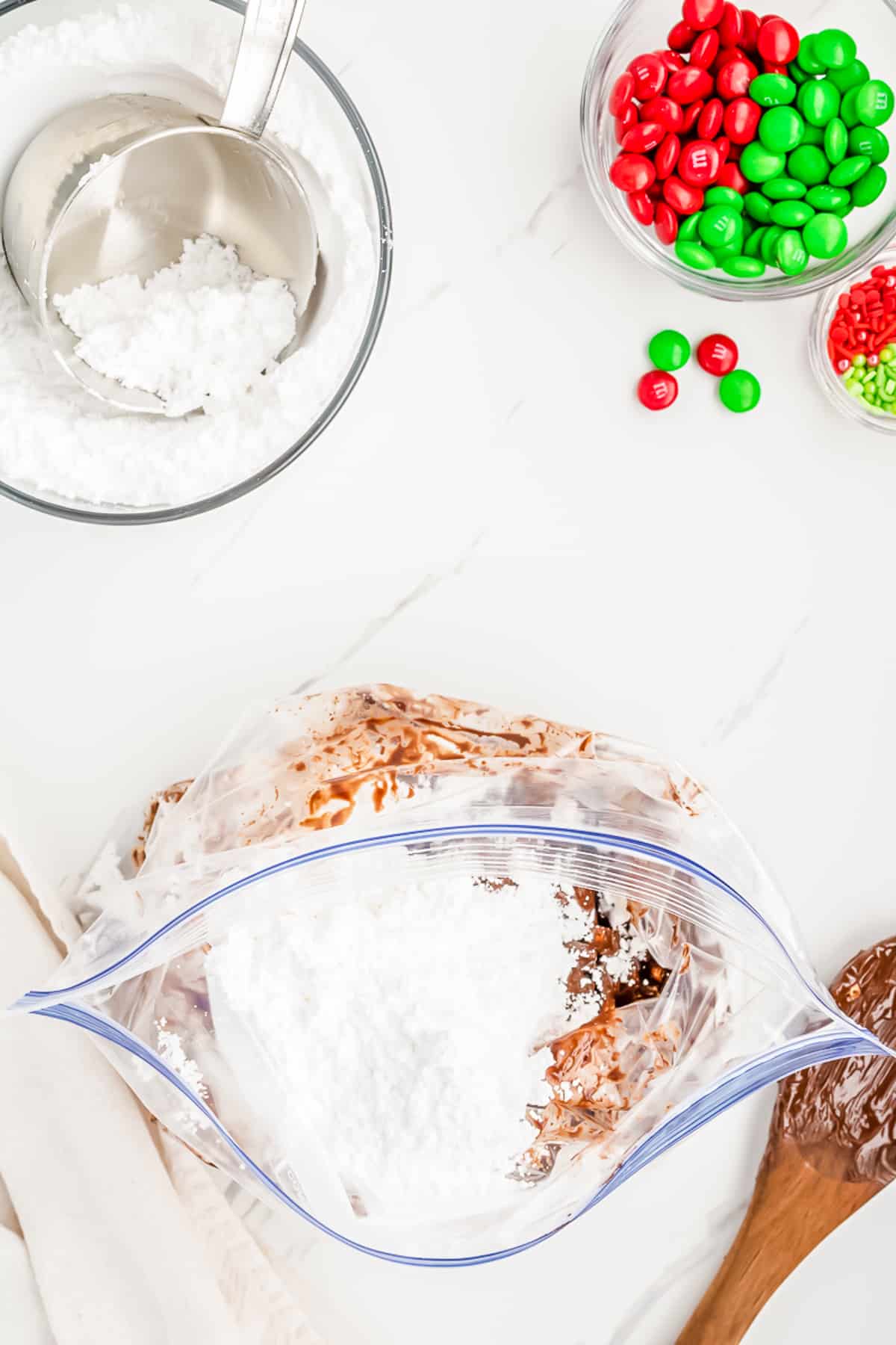 Adding powdered sugar to a chocolate coated cereal in a zip top bag from overhead on a counter with M&Ms and sprinkles in a bowl nearby.