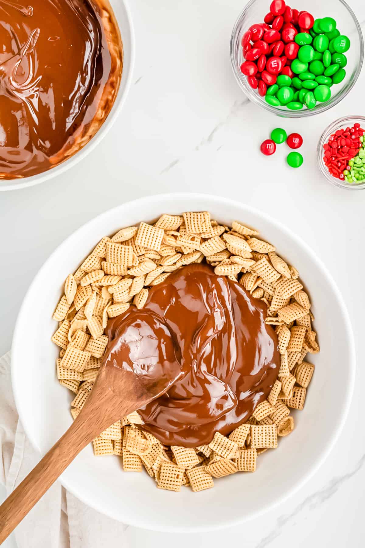 Half of the chocolate sauce being mixed into Chex cereal in a bowl on a counter with more chocolate sauce, M&Ms, and sprinkles nearby on the counter.
