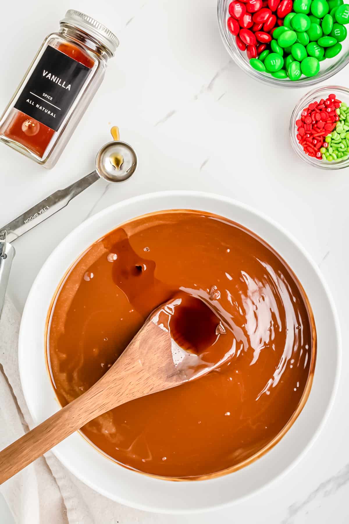 Vanilla being stirred on a wooden spoon into a chocolate peanut butter sauce in a large bowl from overhead with the vanilla bottle,  M&Ms and sprinkles in containers nearby.