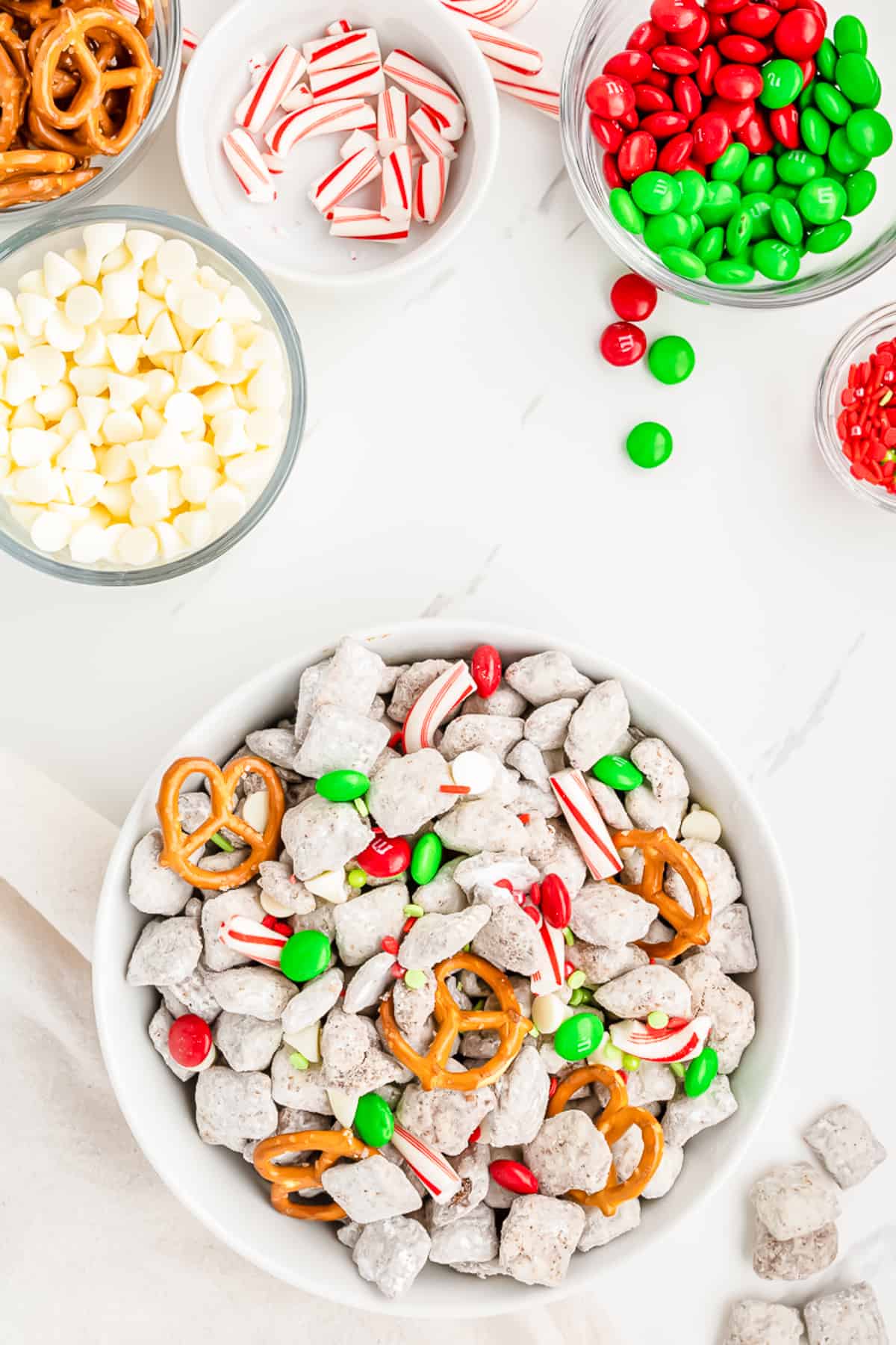 Christmas reindeer chex mix topped with colorful M&Ms, peppermint pieces, white chocolate chips, and pretzels in a bowl on the counter from overhead with the other ingredients in bowls on the counter nearby.