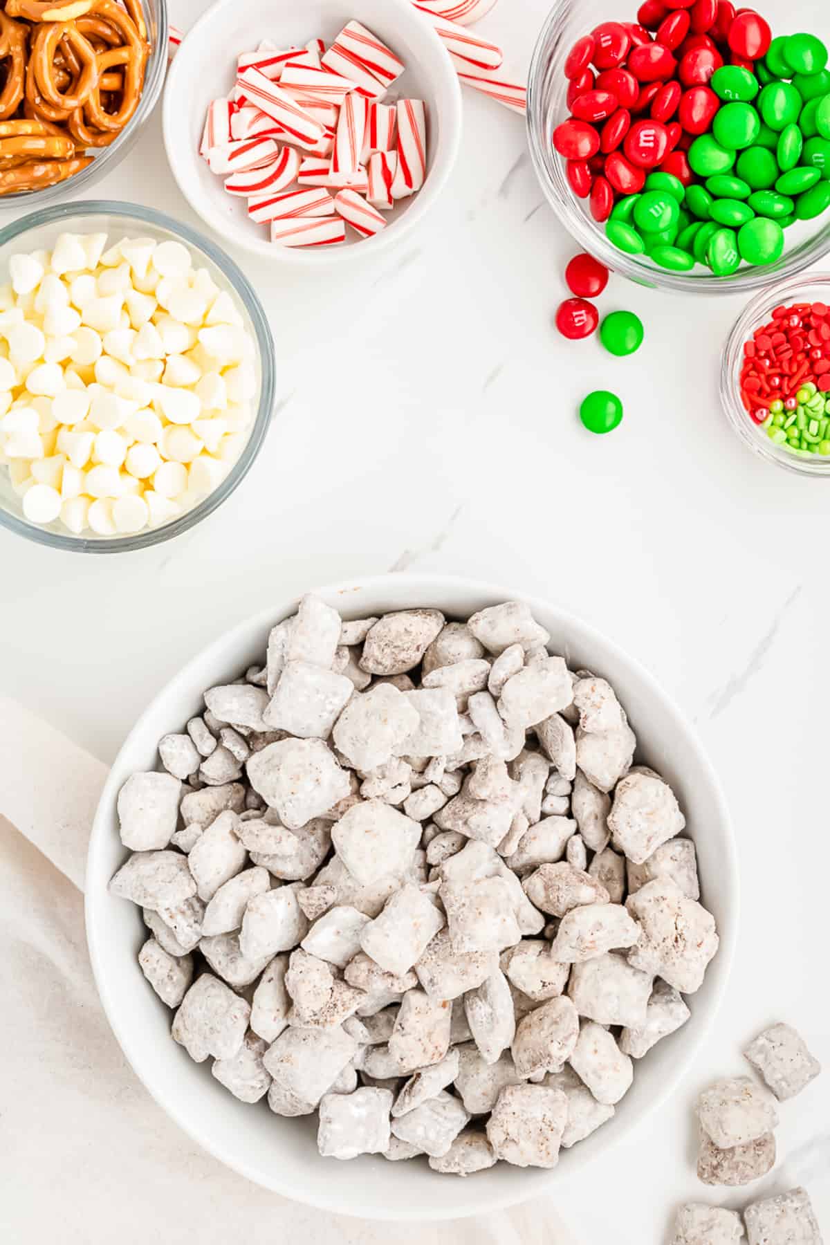 Powdered sugar coated cereal in a large bowl from overhead on a counter with M&Ms, peppermint candy cane pieces, white chocolate chips and pretzels in bowls on the counter nearby.