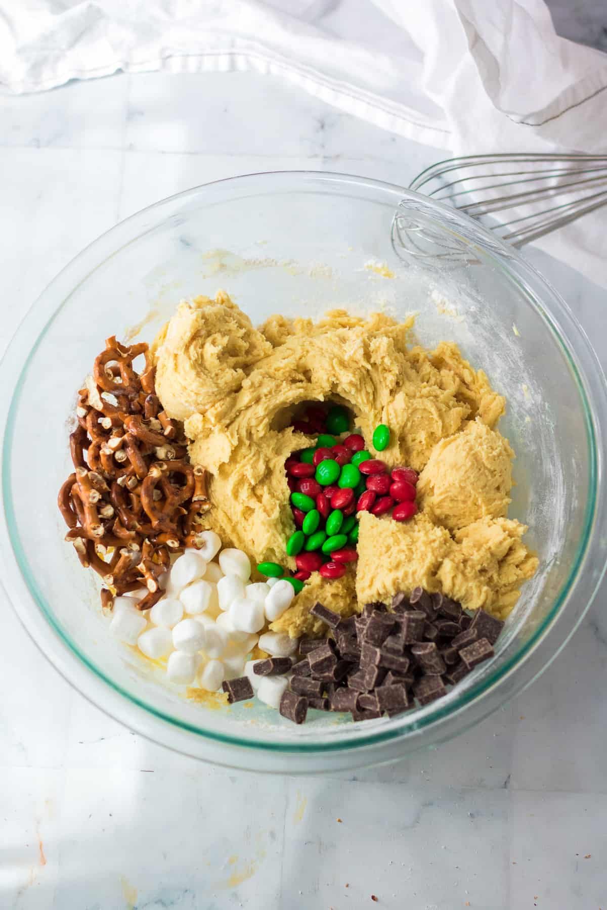 Cookie dough being mixed with red and green M&Ms, chocolate chunks, pretzels and marshmallows in a mixing bowl from overhead on a counter.