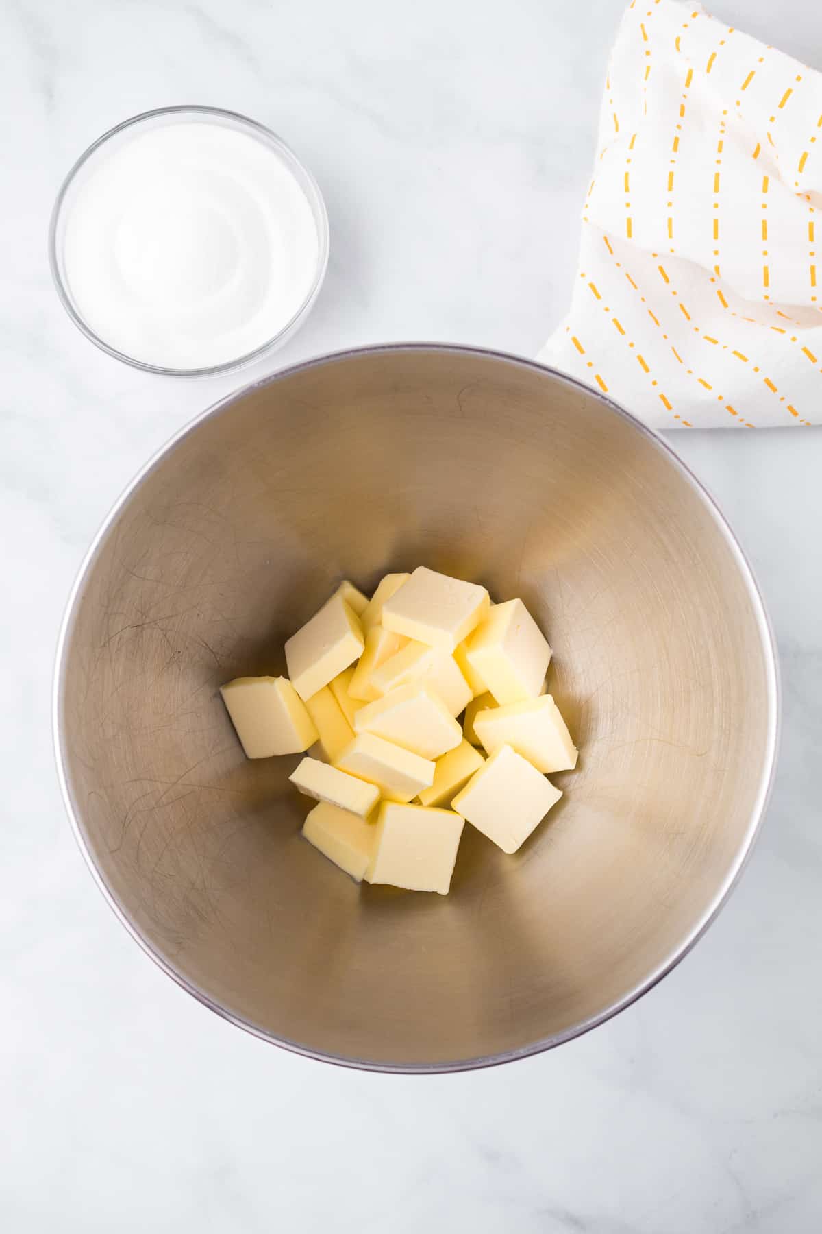 Cubes of butter in the base of a stand mixer next to a bowl of sugar on a counter from overhead.