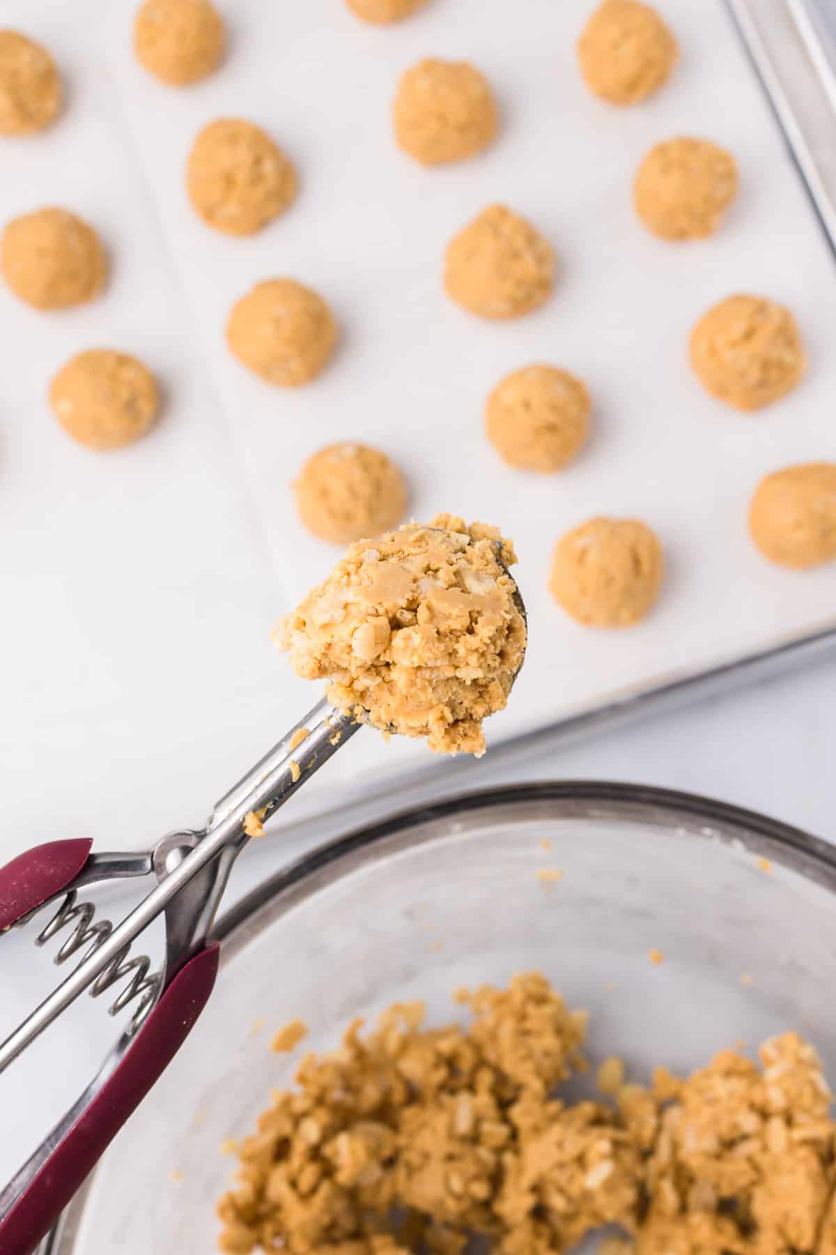 A cookie scoop scoops peanut butter rice krispie dough from a bowl from overhead on a counter while more peanut butter balls sit on a pan nearby evenly spaced from overhead..