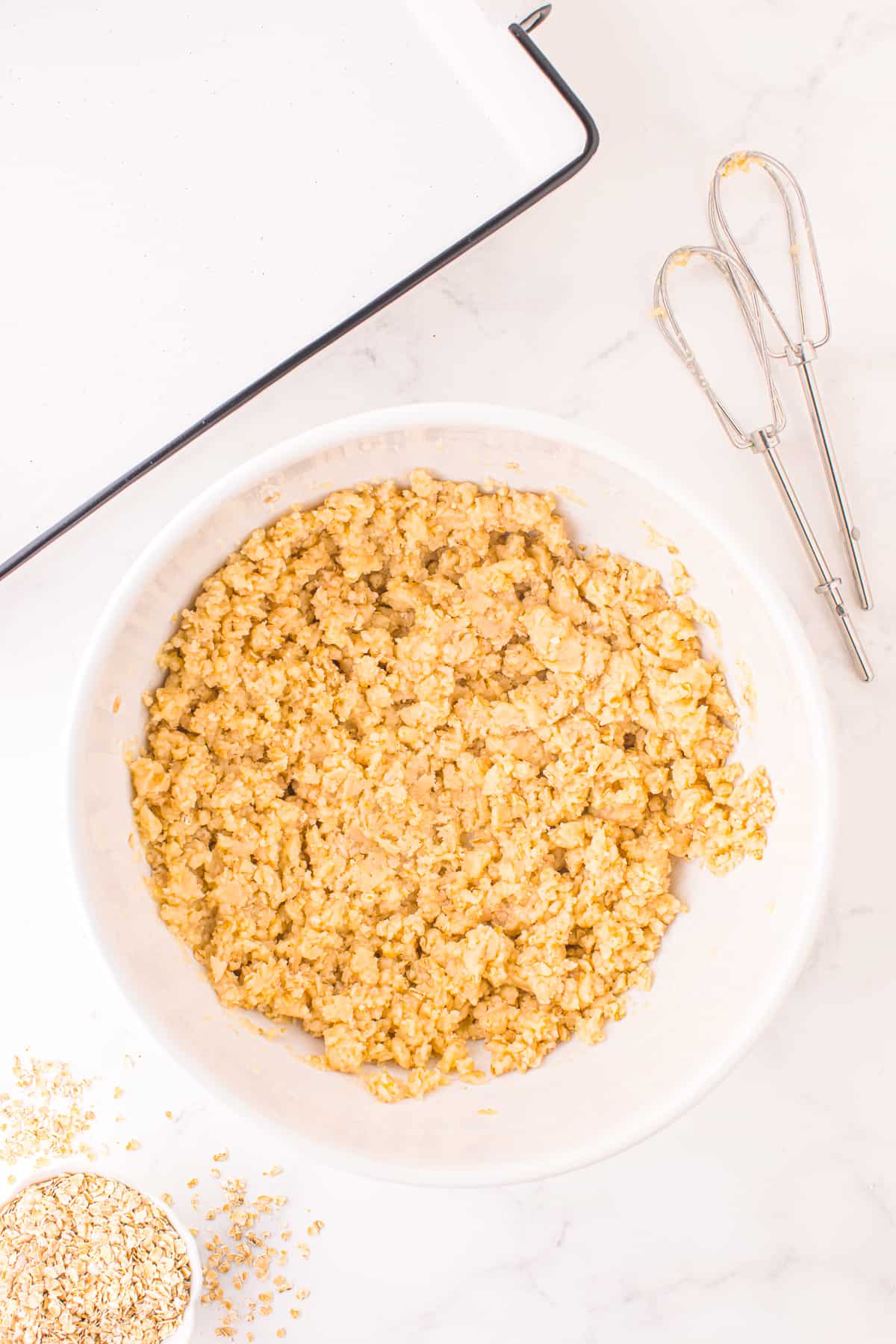 Dough for cranberry crumble bars mixed in a large mixing bowl on a counter with a thick shaggy apperance, with mixer beaters and a large pan on the counter nearby.