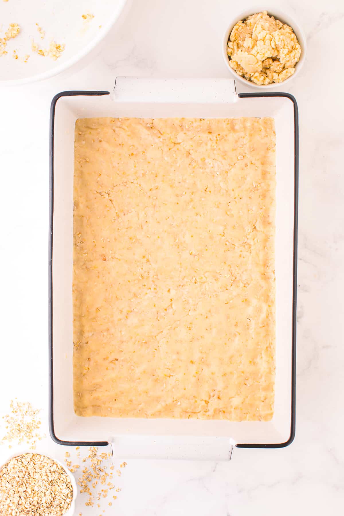 A buttery dough is pressed into the bottom of a large rectangular pan from overhead with more dough in a small bowl on the counter nearby.