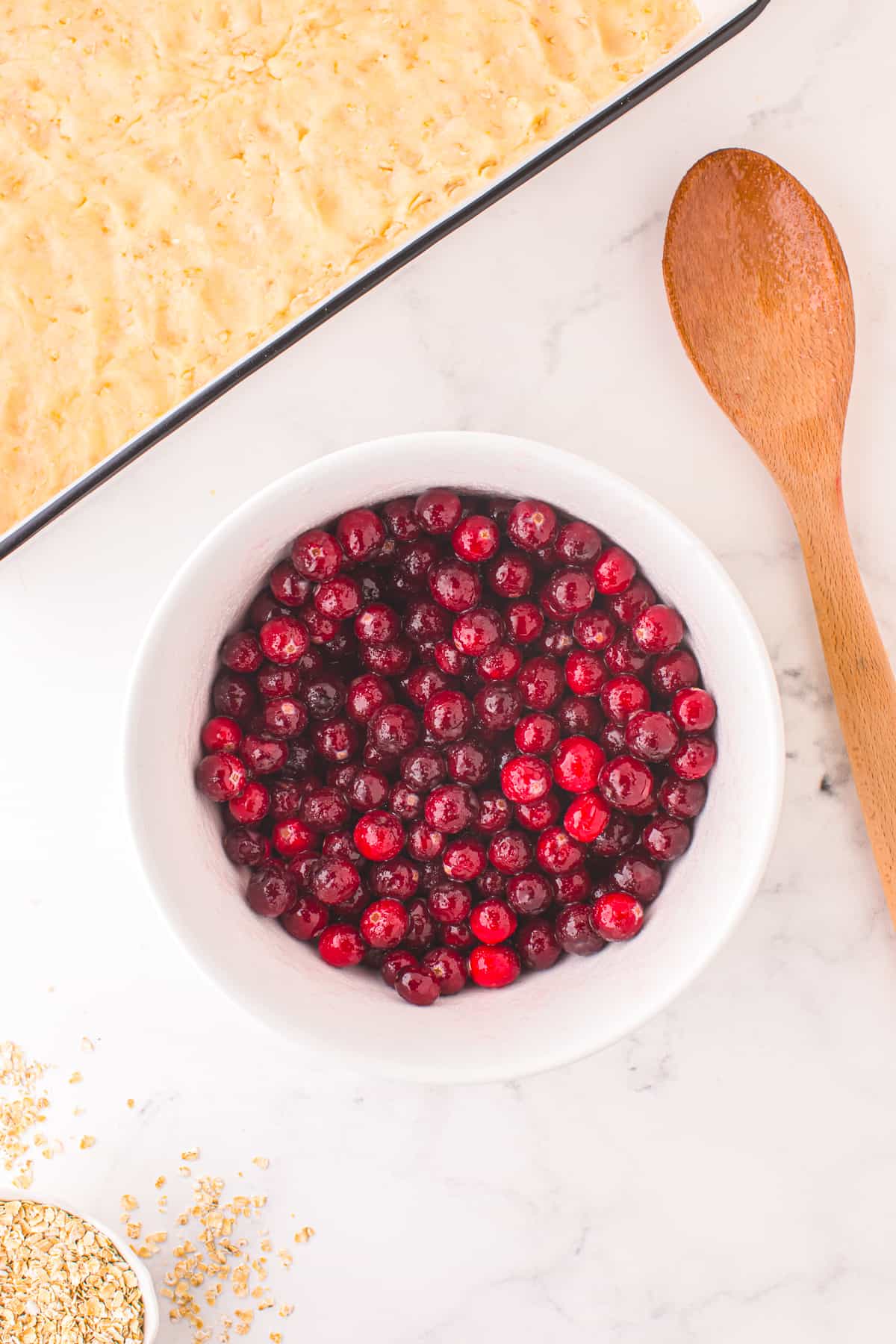 Cranberries in a large bowl mixed with other liquid ingredients from overhead on a counter with a wooden spoon and a large pan with the dough pressed into the bottom on the counter nearby.