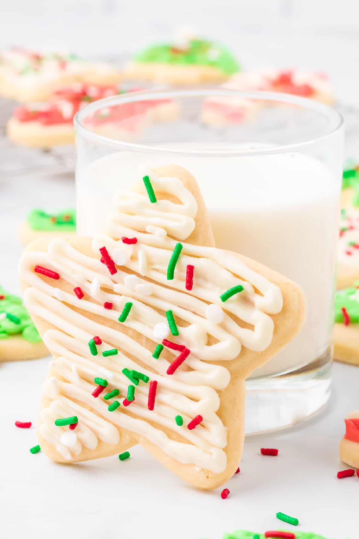 Frosted sugar cookie with red, green and white sprinkles and frosting leaning on a glass of milk with more Christmas cookies on the counter nearby.