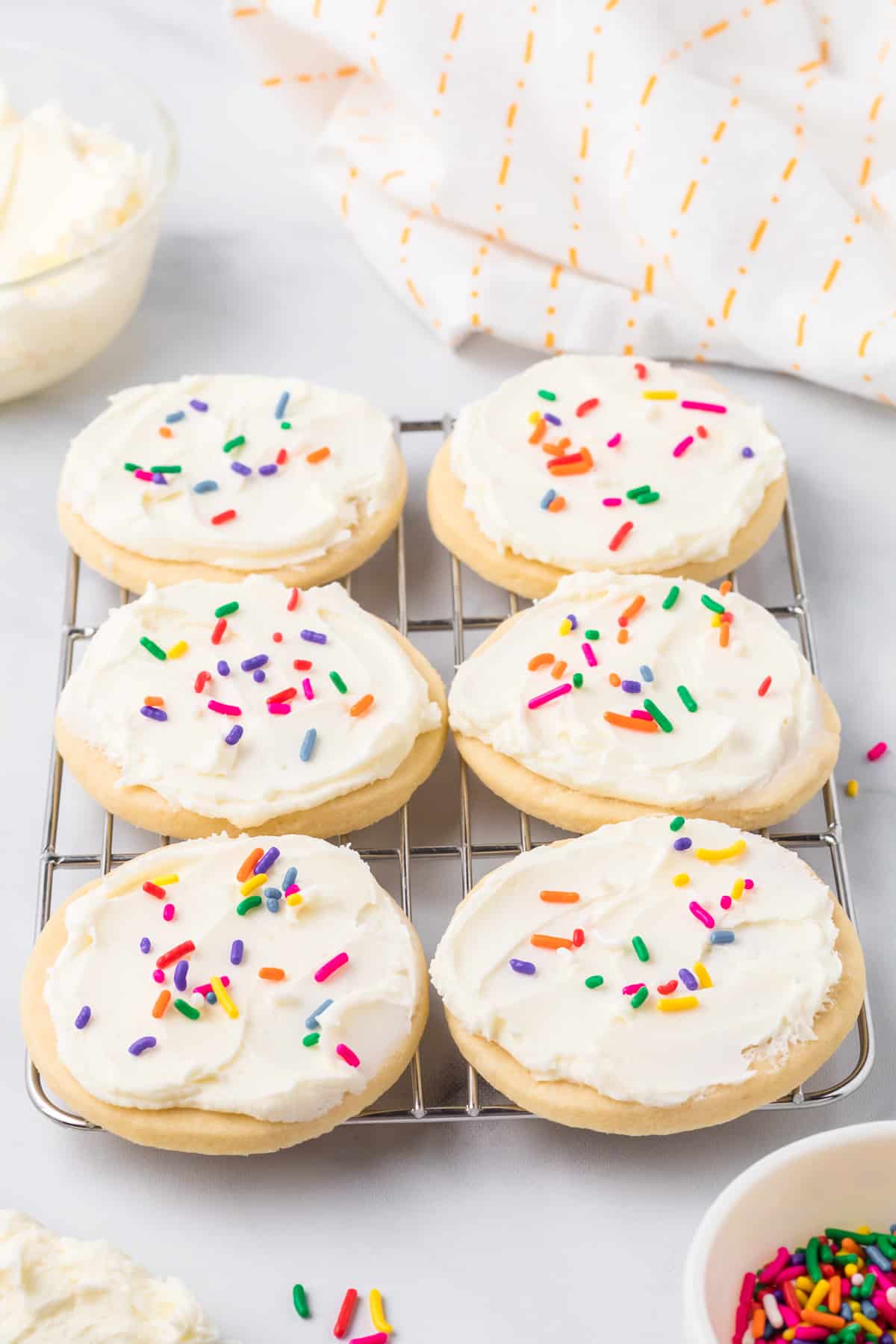 Round sugar cookies frosted with white frosting and rainbow sprinkles on a wire rack on a counter from the side.