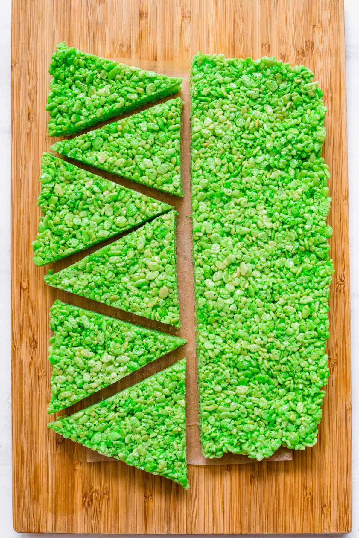 Large slice of green rice krispie treat cut into two long pieces with one piece cut into six equal triangles on a cutting board from overhead.