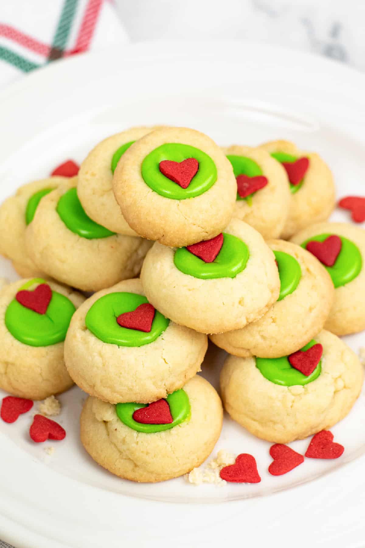 Grinch thumbprint cookies piled high on a platter with red sprinkles in the middle of each green thumbprint center from the side on a counter.