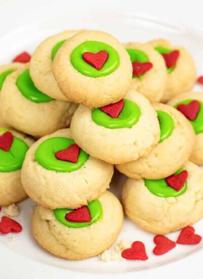 Close up of a stack of thumbprint cookies with a grinch bright green center with a heart sprinkle on a plate on a counter.