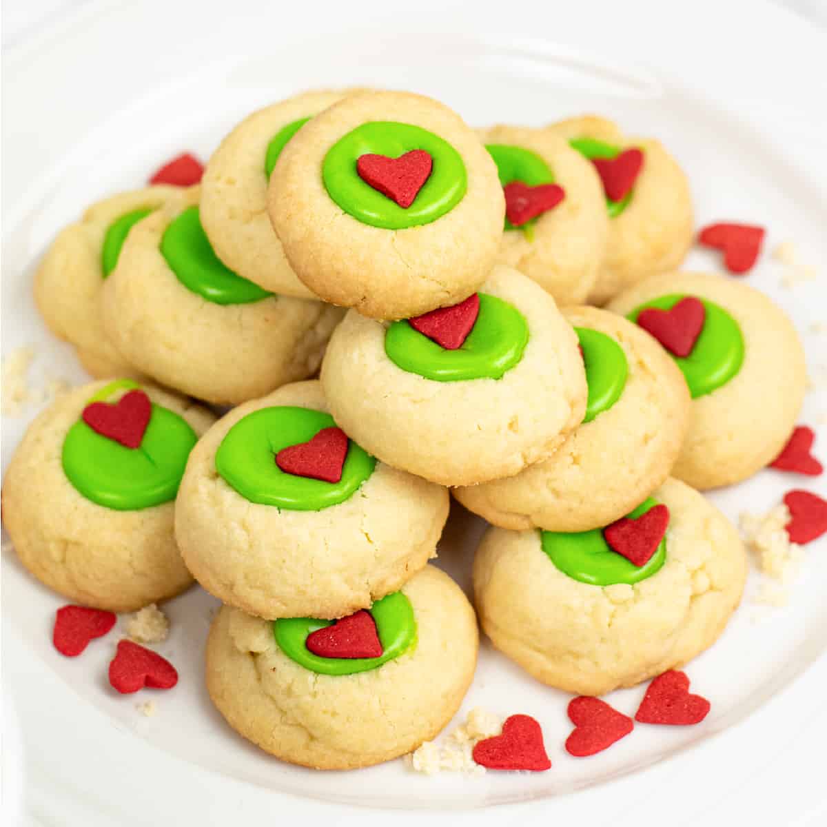 Close up of a stack of thumbprint cookies with a grinch bright green center with a heart sprinkle on a plate on a counter.