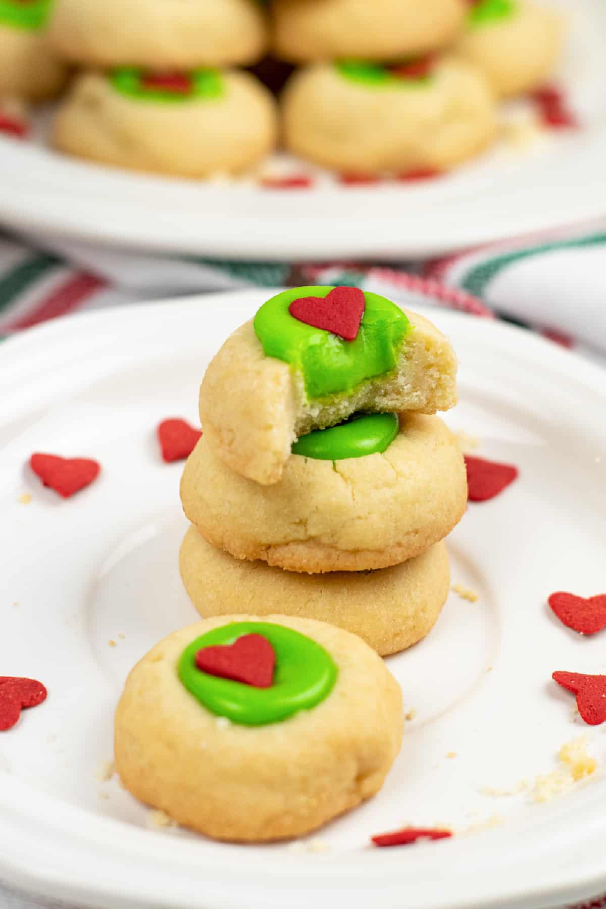 Four thumbprint cookies stacked on a plate with the top cookie missing a bite into the green thumbprint filling. A plate of more grinch thumbprint cookies sits behind on a plate on the counter.