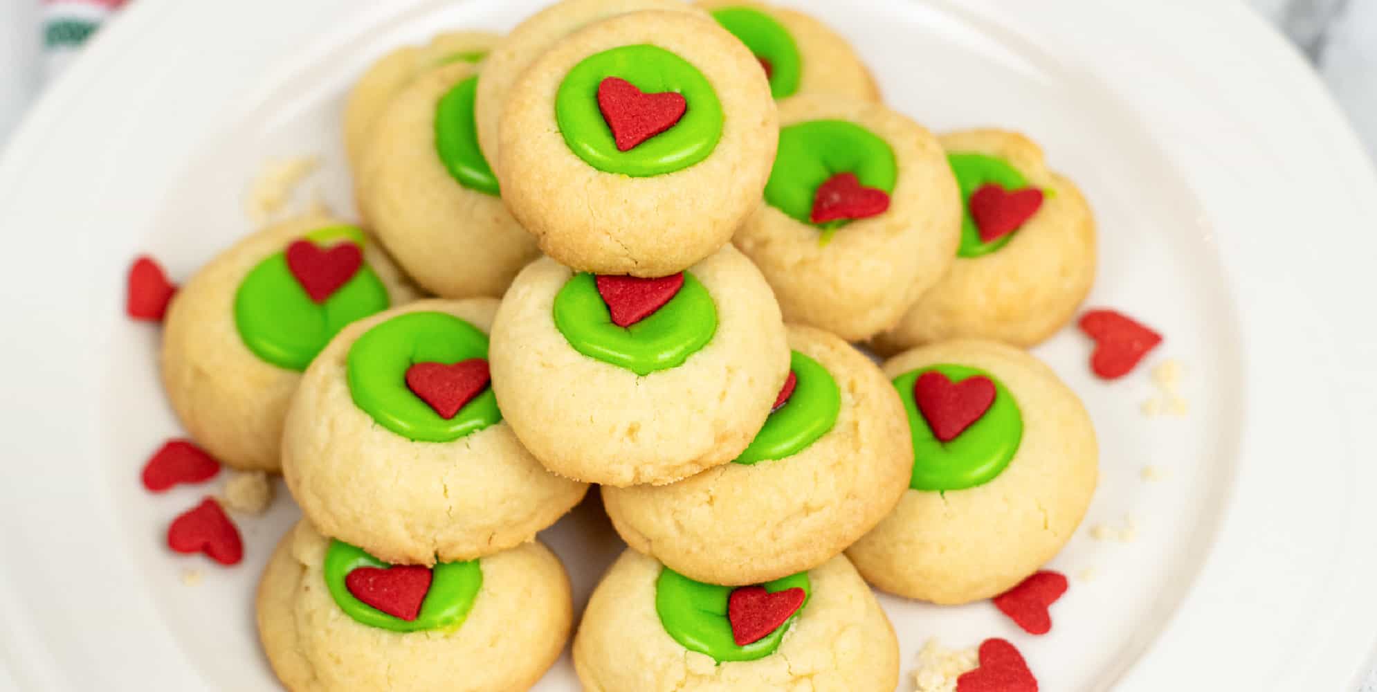 Wide view of a stack of thumbprint cookies full of green filling with a red heart sprinkle in the middle of each cookie stacked on a plate high on a counter.