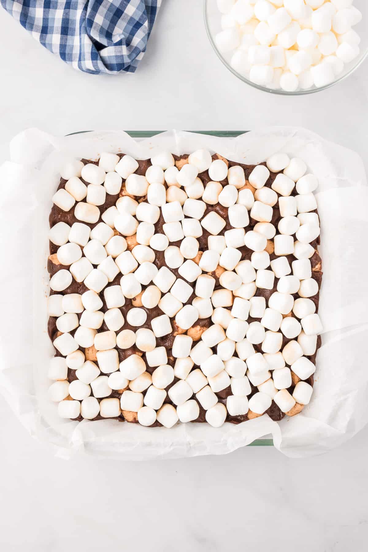 Mini marshmallows covering a pan full of hot chocolate fudge with more marshmallows in a bowl nearby.