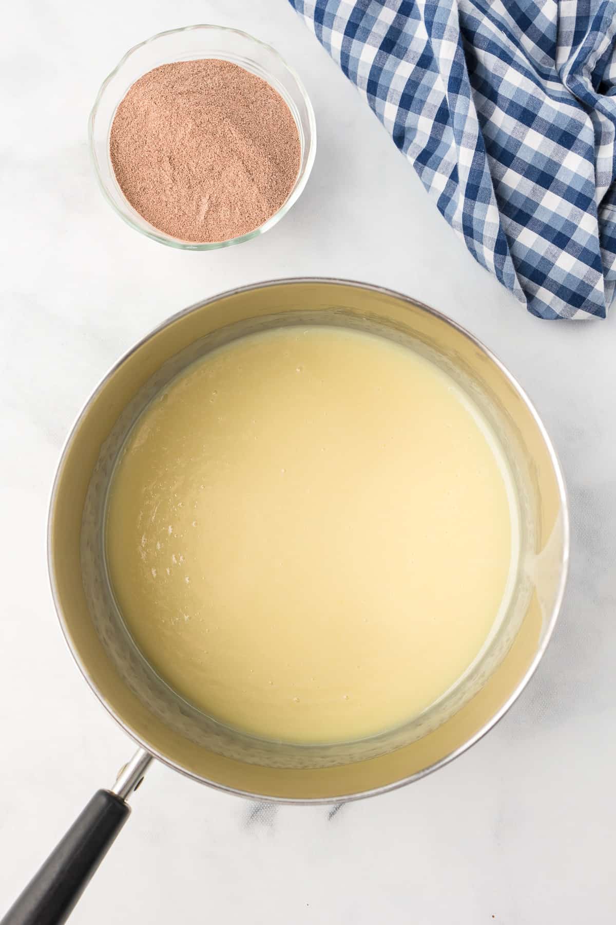 Sweetened condensed milk mixture in a pan from above with hot cocoa mix in a bowl nearby on the counter.