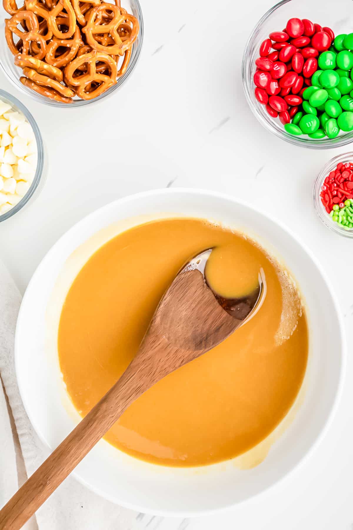 Melted peanut butter sauce in a bowl with a wooden spoon from overhead on a counter with other ingredients nearby in bowls from overhead.